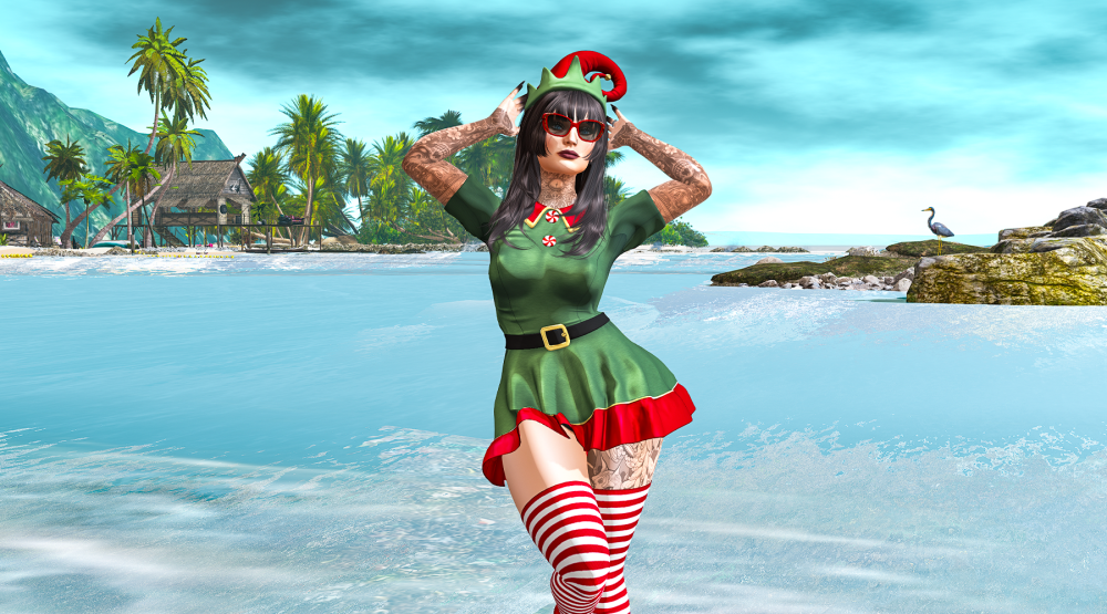 A stylish female avatar dressed in a festive green and red elf outfit with striped stockings poses confidently on a tropical beach in Second Life. Behind her are palm trees, stilted beach huts, and a calm blue sea under a bright sky — a playful clash of Christmas spirit and summer vibes.