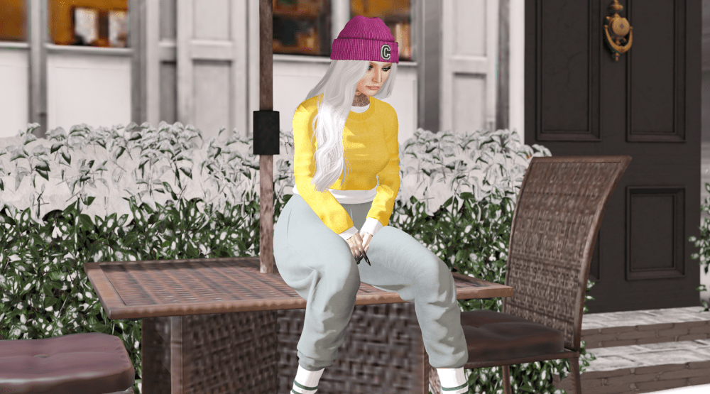 A Second Life avatar with long white hair sits outdoors at a patio table, wearing a yellow sweater, light grey sweatpants, white socks, and a magenta beanie with the letter “C.” The avatar appears relaxed and thoughtful, surrounded by greenery and a building entrance in the background.